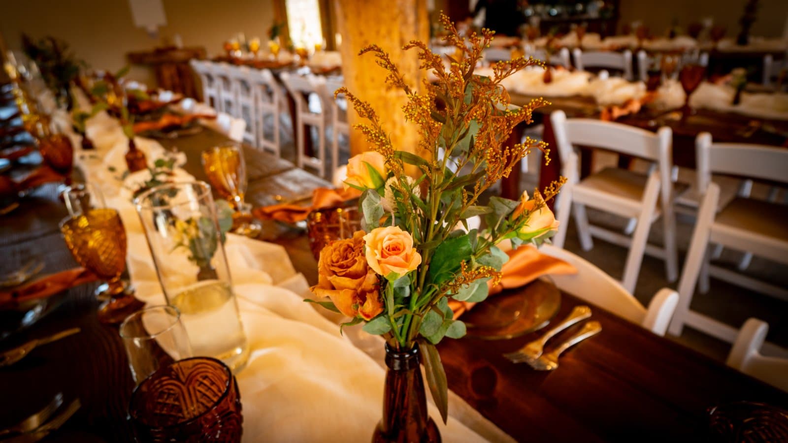 Amber bud vase of roses and goldenrod on a cheesecloth runner, tables receding into the warm barn interior