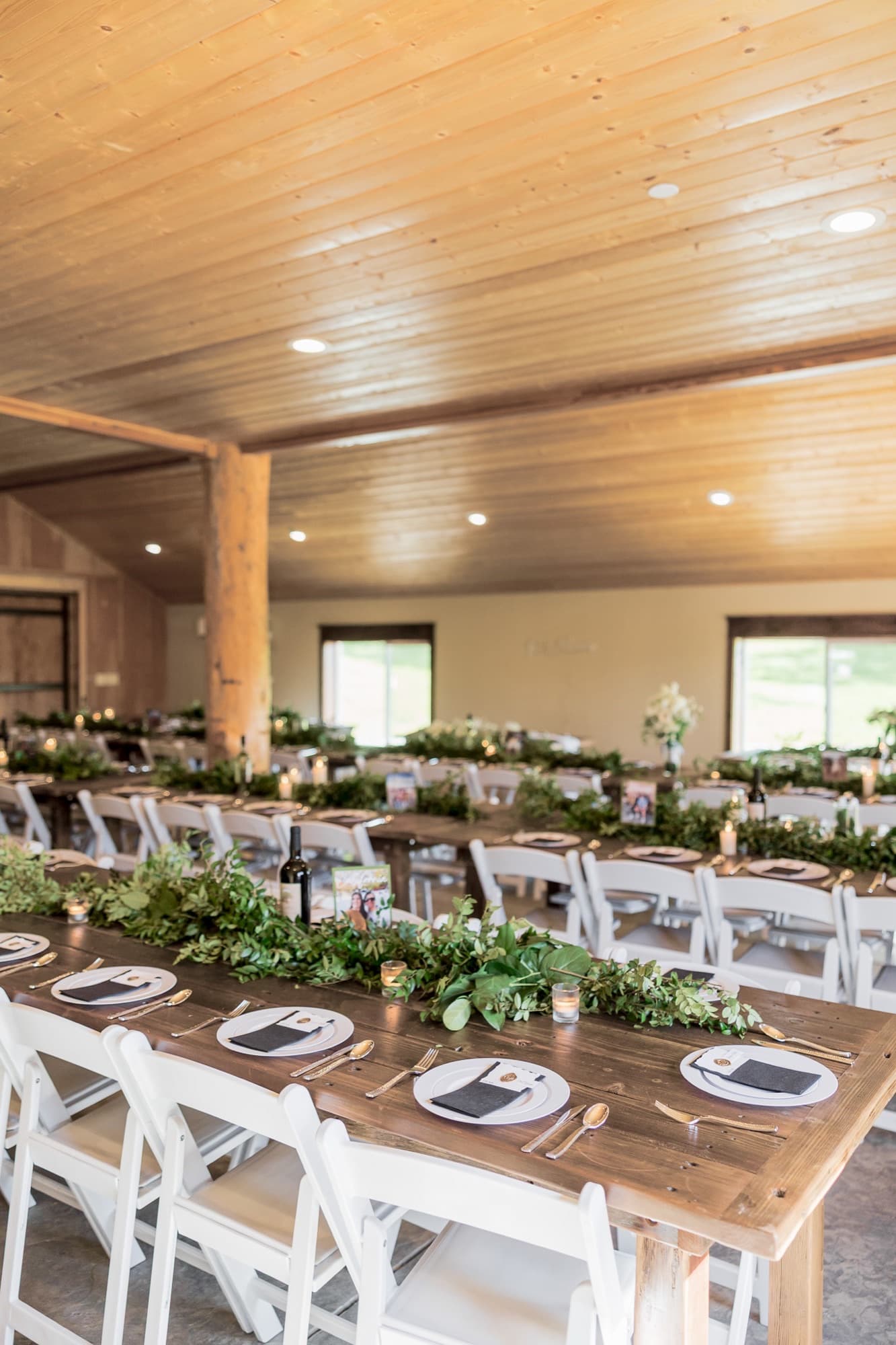 Fresh greenery garlands and candles down every farm table at a winery wedding in the barn, wine bottles set between them