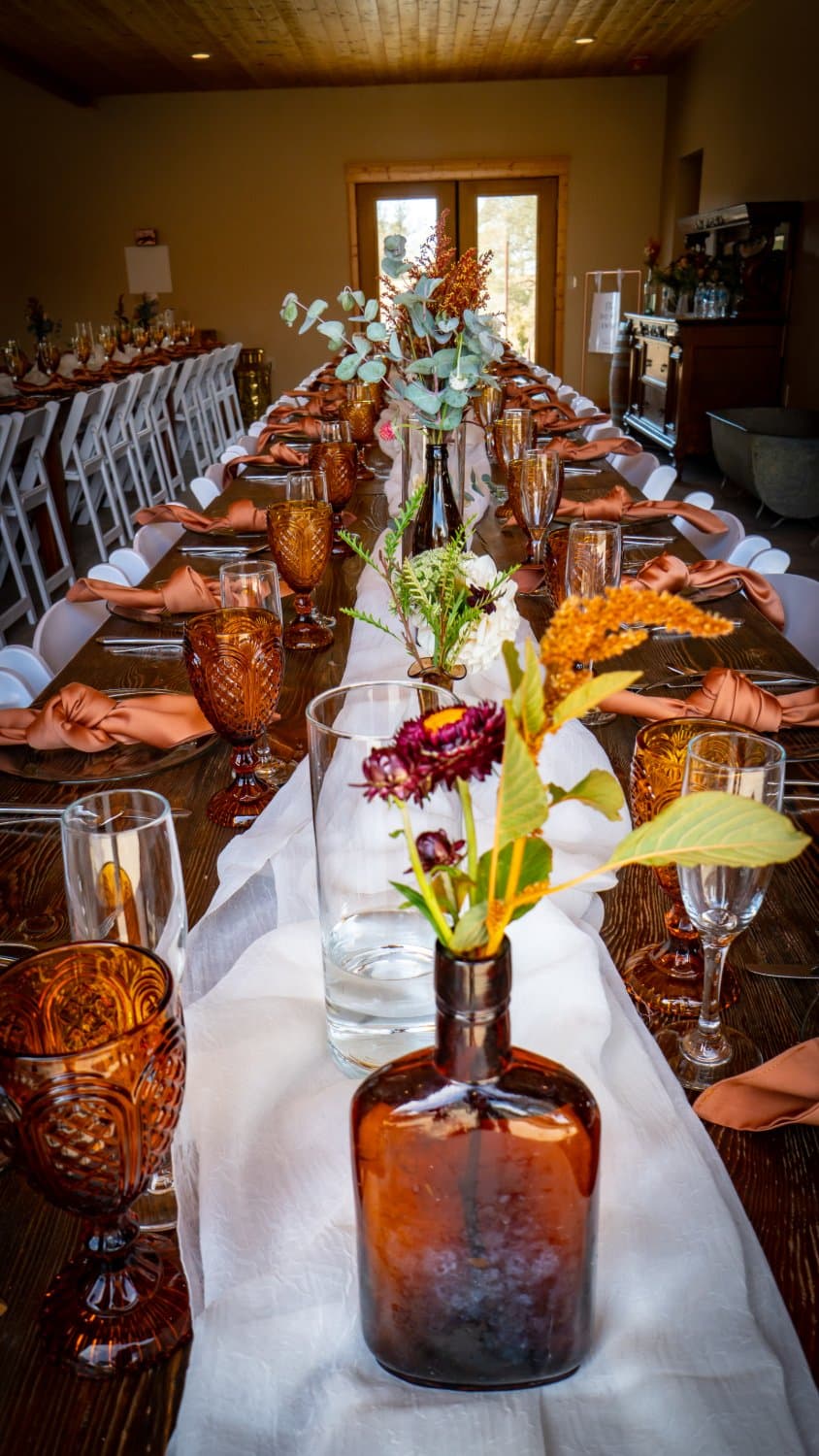 View down a long farm table set with amber goblets, rust napkins and bud vases toward the barn's west doors