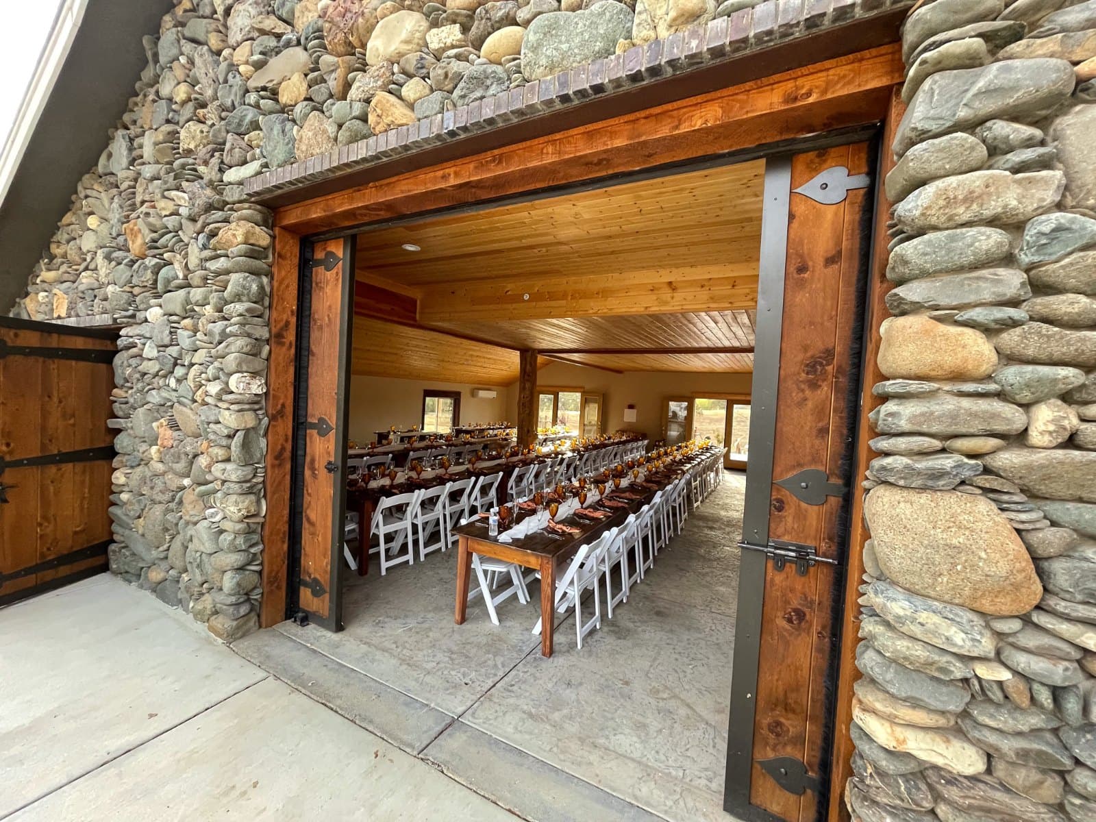 Long farm tables set with amber glassware and white folding chairs seen through the open stone barn doors