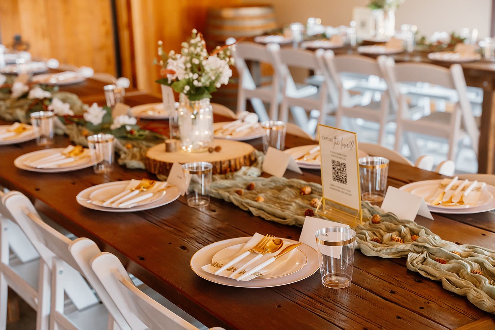 Long farm table with a sage cheesecloth runner, gold flatware, wood-slab centerpiece and a fairy-light mason jar at a rustic wedding