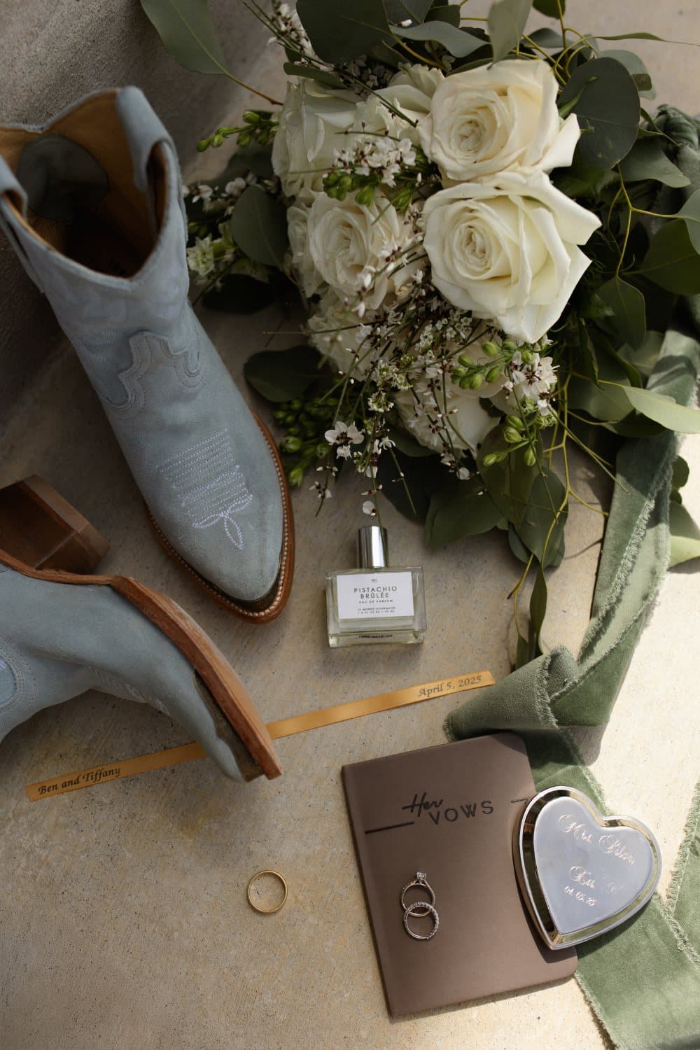 Pale blue suede cowboy boots beside rings, vow book and a white rose bouquet on the barn concrete