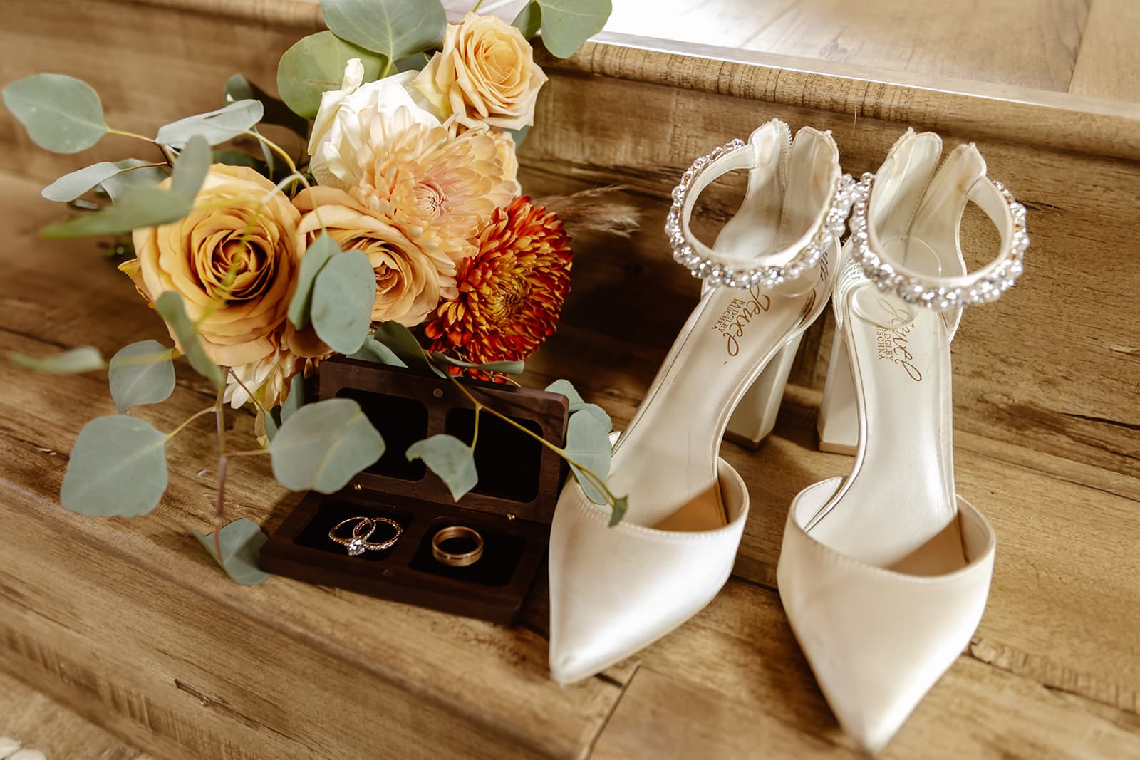 Jeweled bridal heels beside a walnut ring box and apricot florals on the barn's wooden stairs
