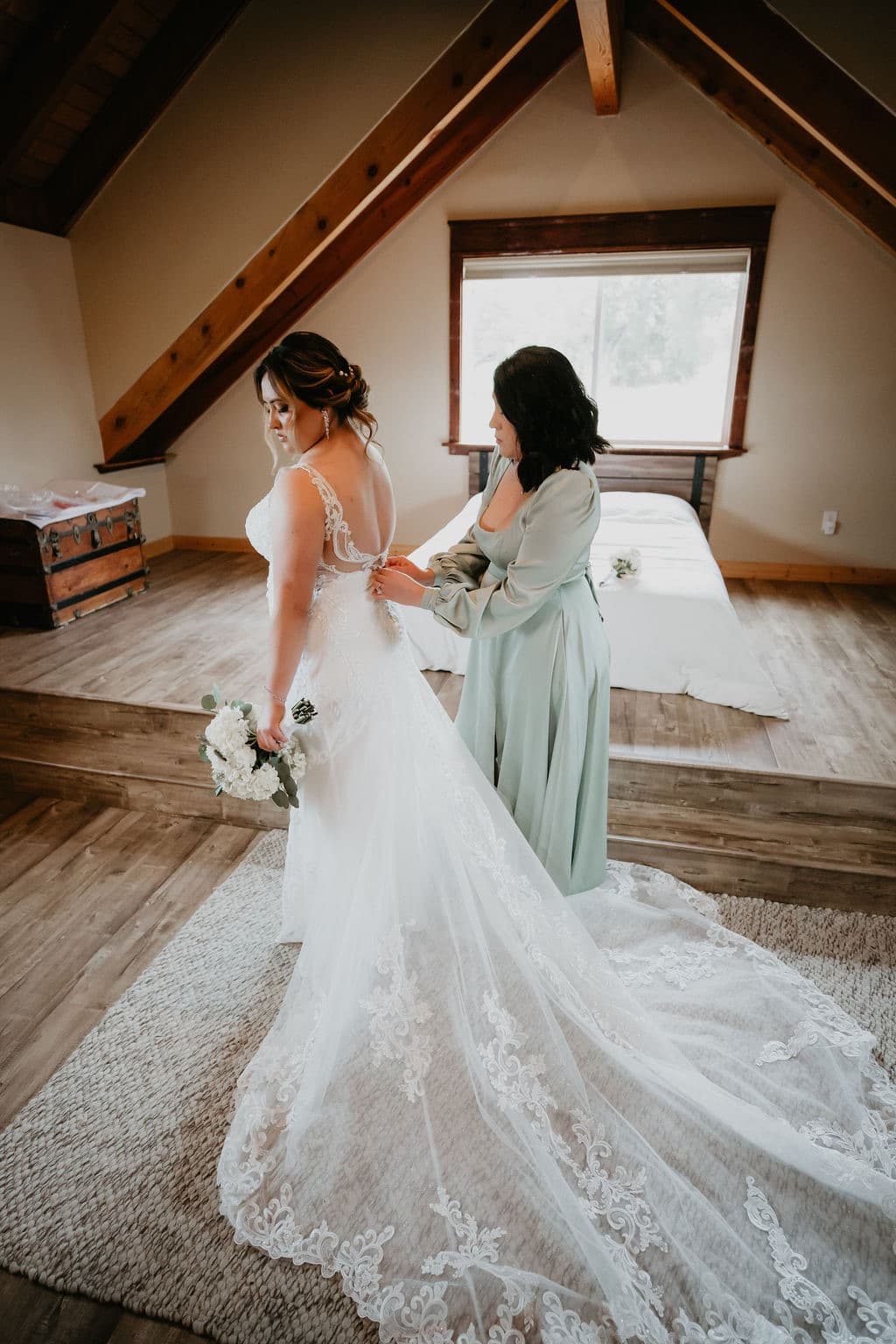 Bridesmaid in sage buttons the bride's lace gown under the bridal suite's timber beams at a barn wedding