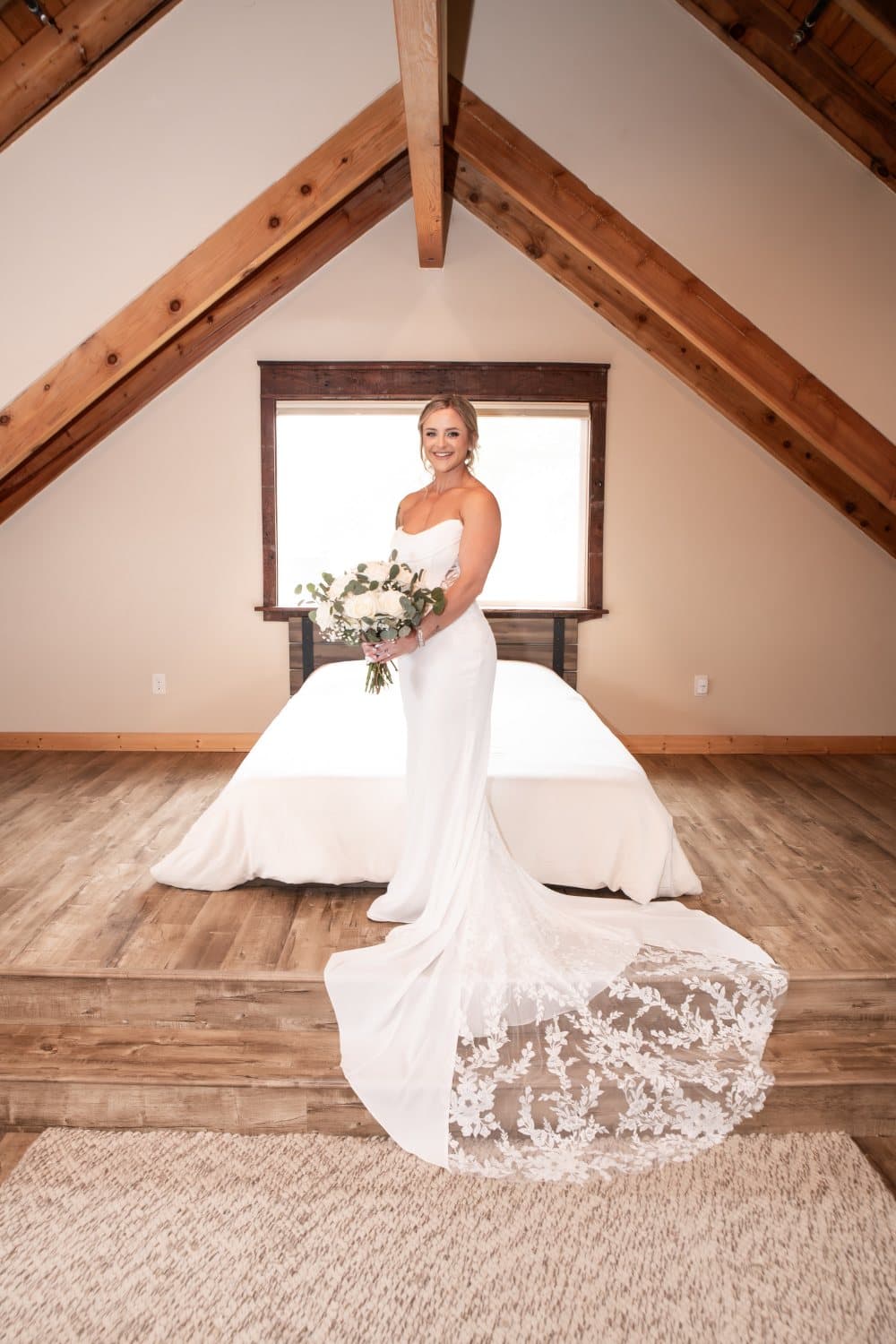 Bride stands under the exposed timber beams of the bridal suite holding a white rose and eucalyptus bouquet