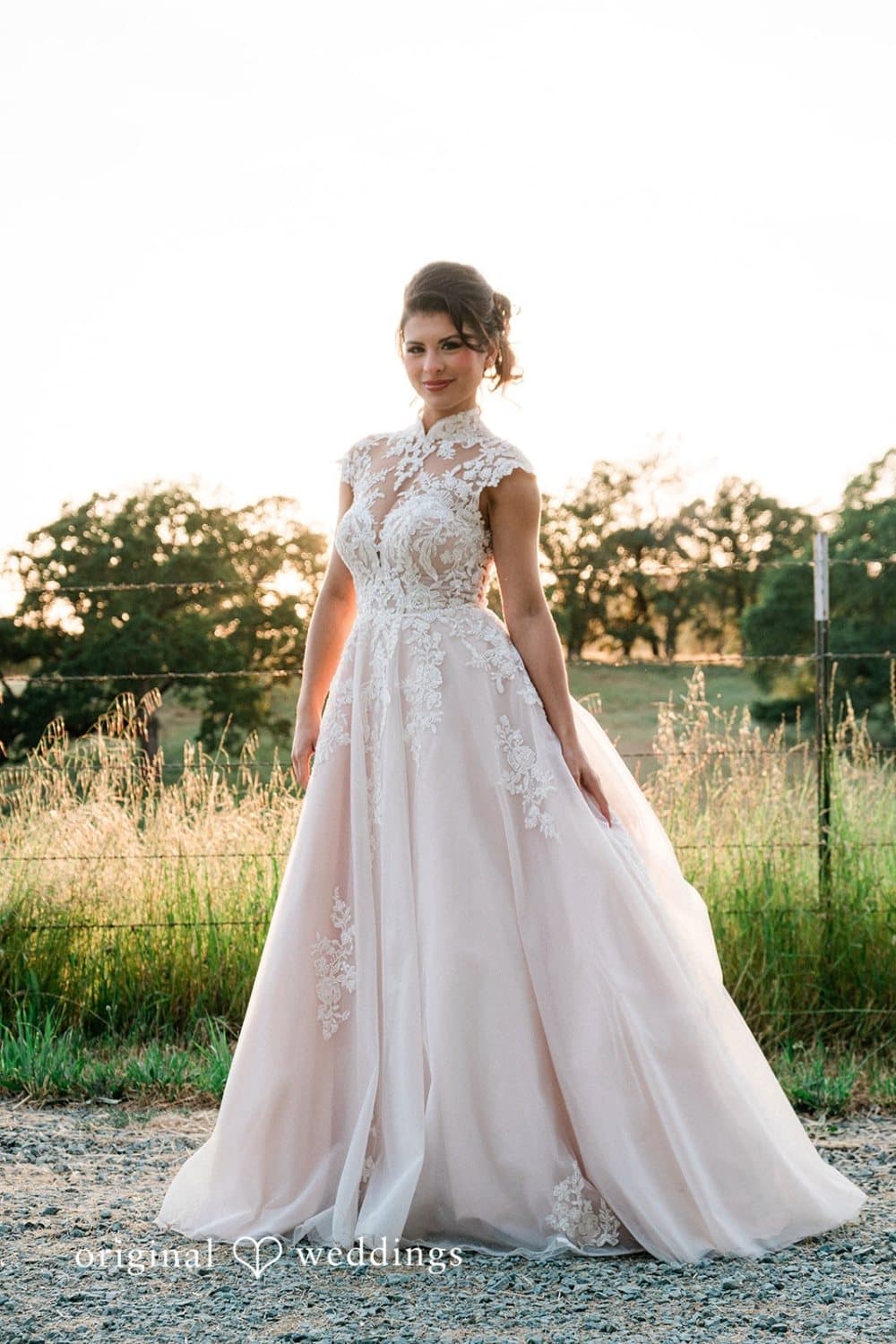 Bride in a blush lace ballgown stands at the ranch fence line with the sun setting through the oaks