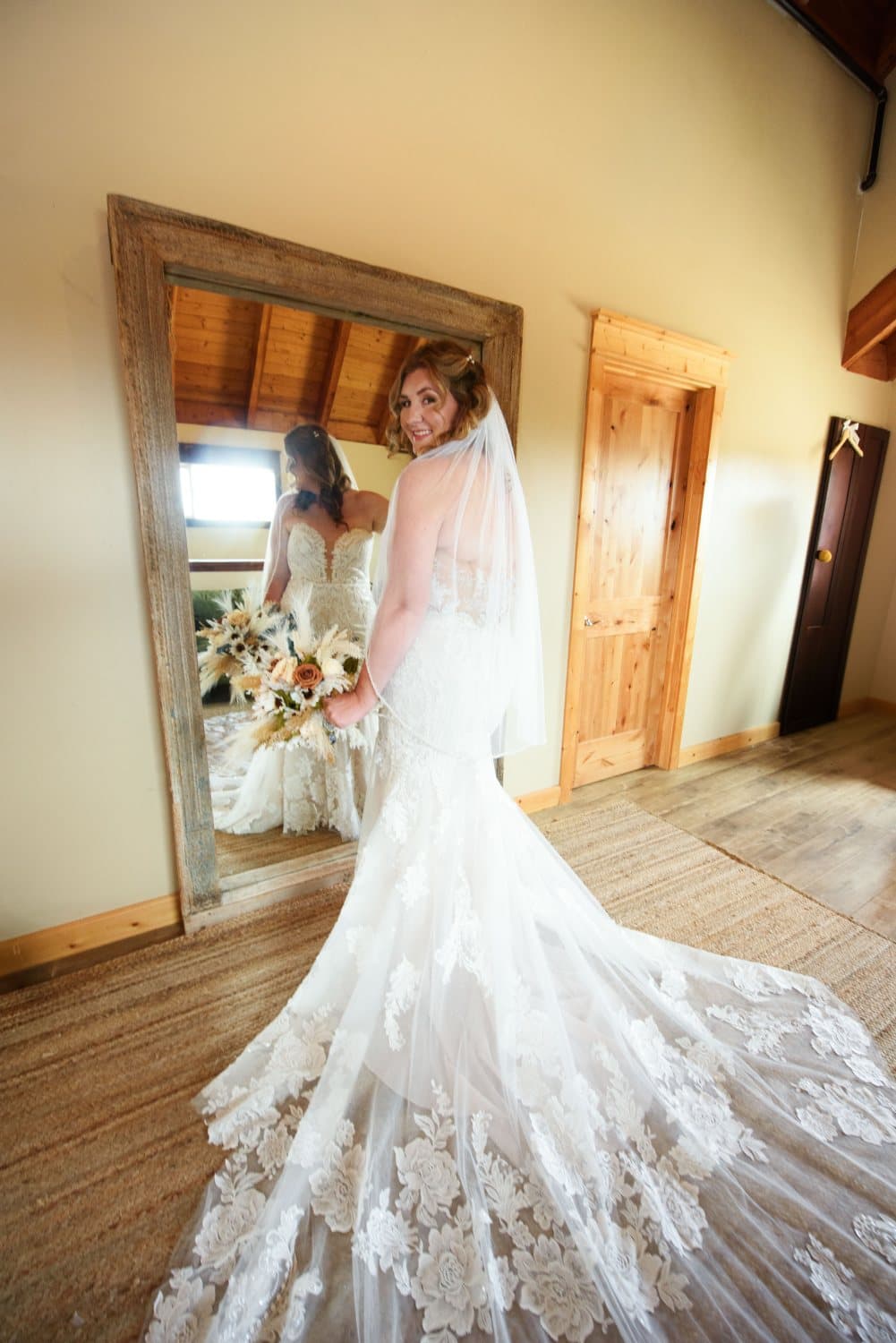 Bride in a lace mermaid gown looks back at the camera by the suite mirror holding a pampas and dried-flower bouquet