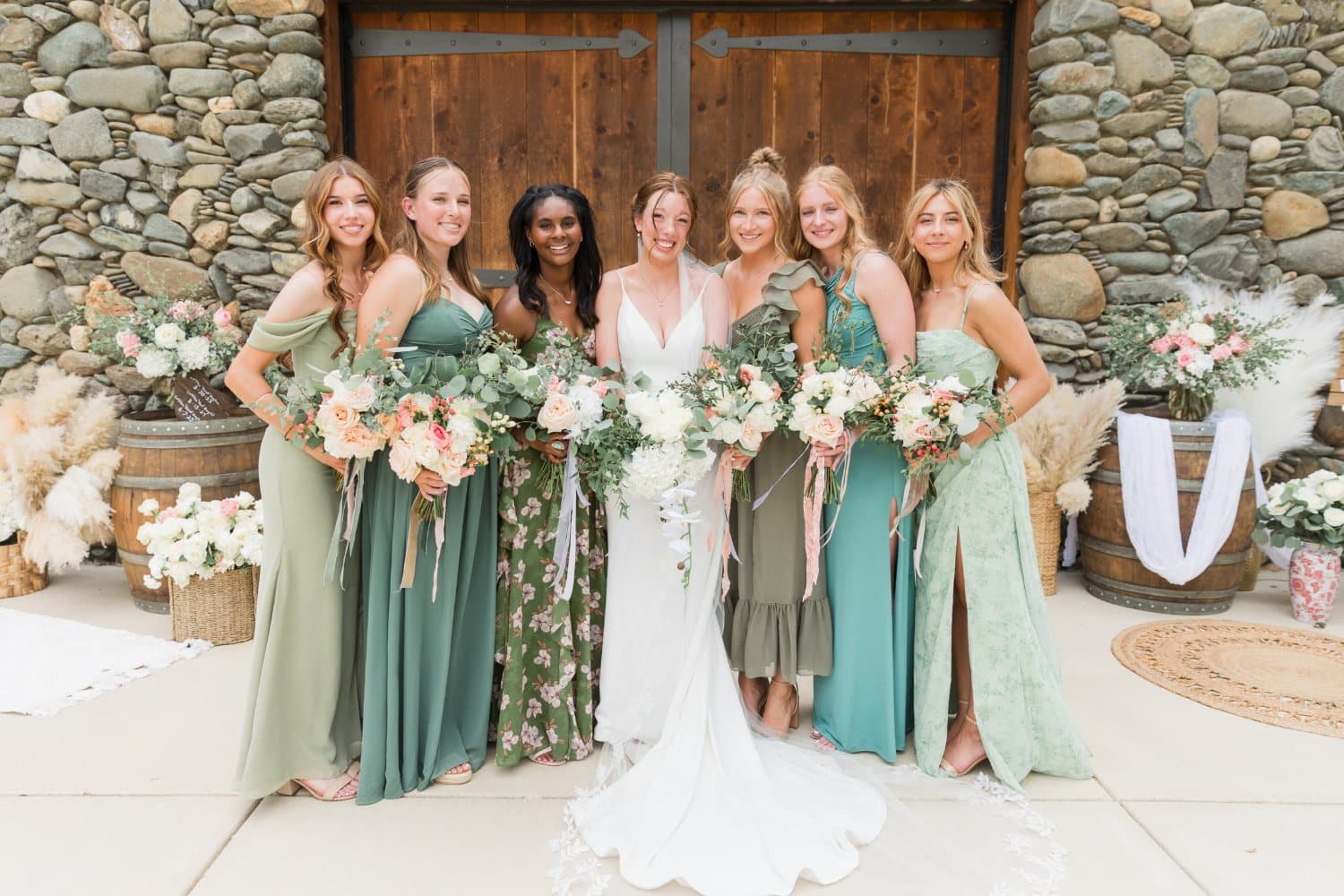 Bridesmaids in mixed green dresses raise their bouquets outside the stone barn doors, framed by wine barrels and pampas grass