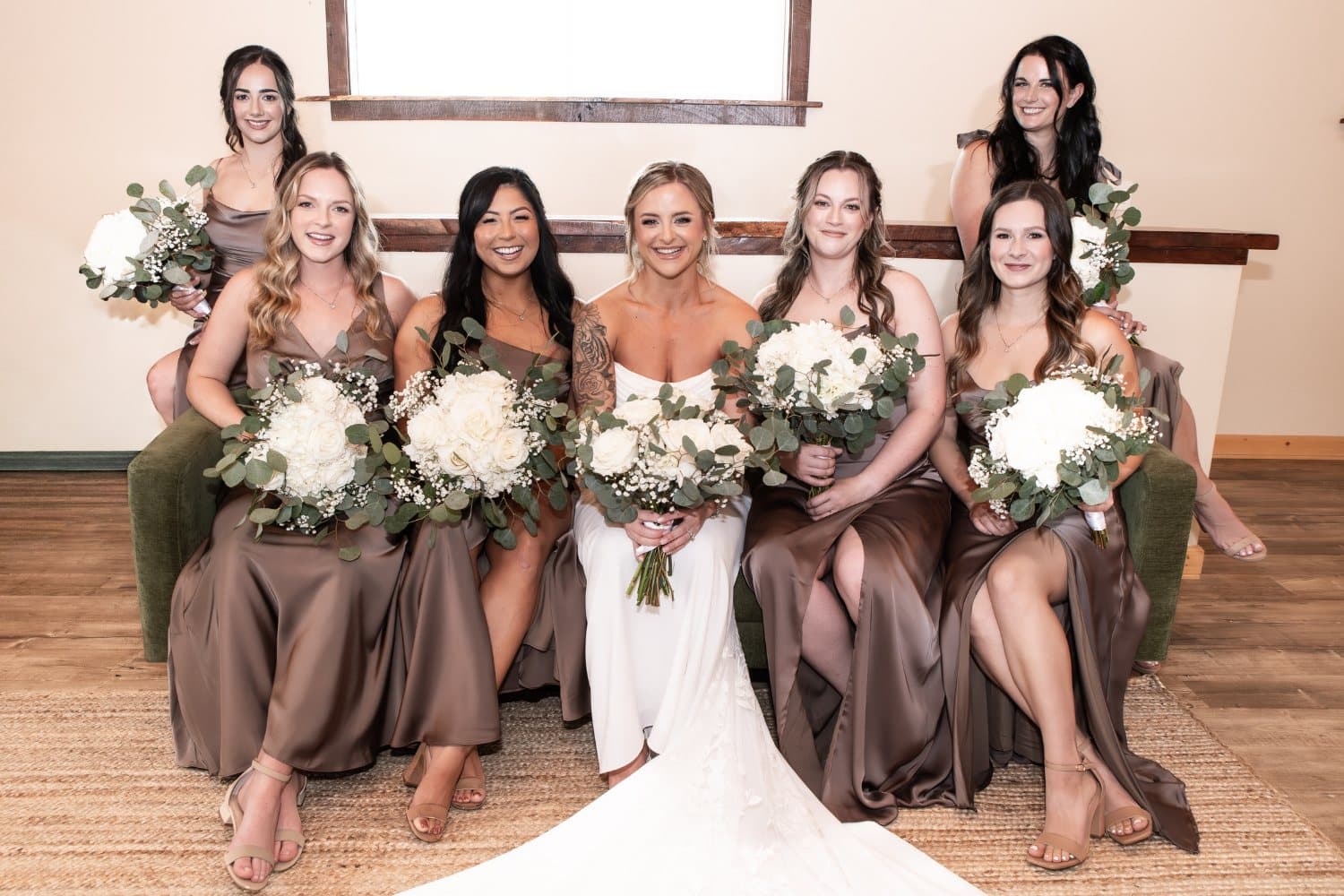Bride and six bridesmaids in mocha satin gowns seated together in the bridal suite with white rose bouquets
