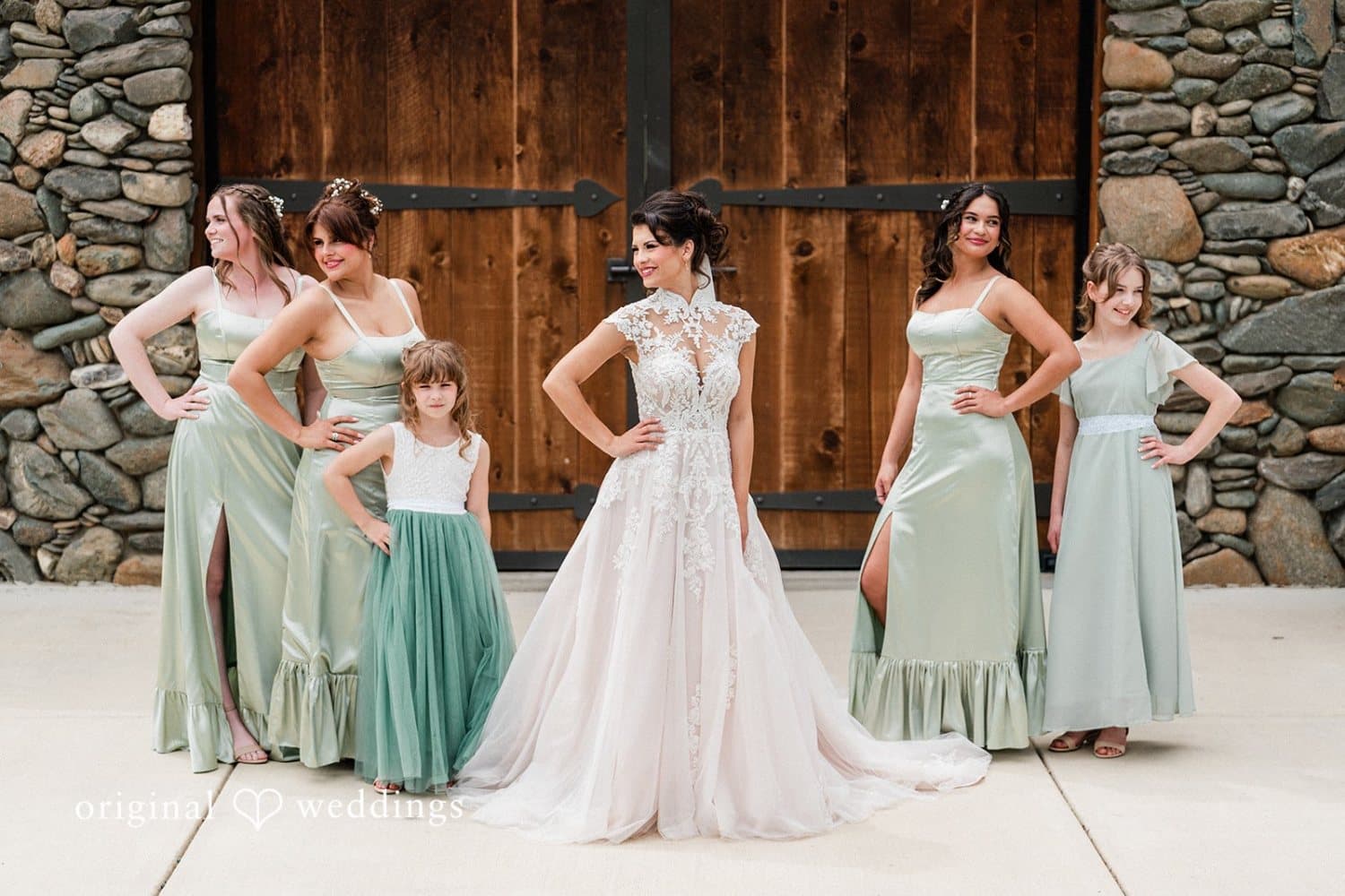 Bride and her bridesmaids in sage green pose hands-on-hips at the stone barn doors with the flower girl