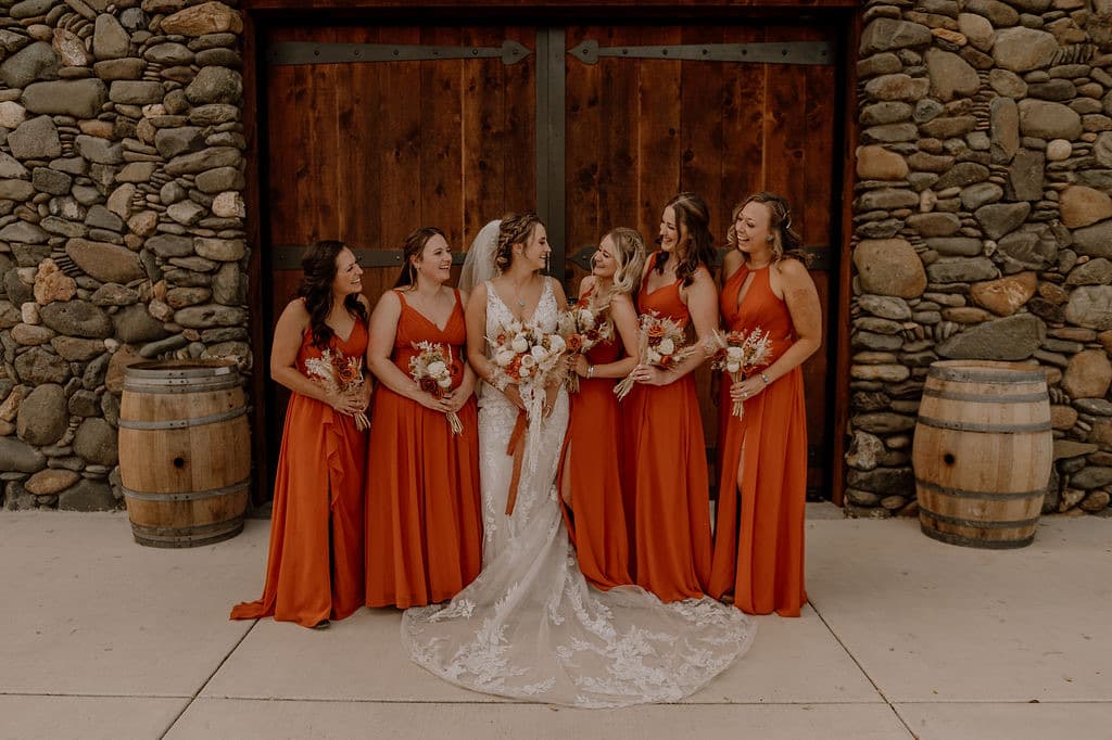Bride laughing with bridesmaids in terracotta gowns at the barn doors, wine barrels either side, winery wedding