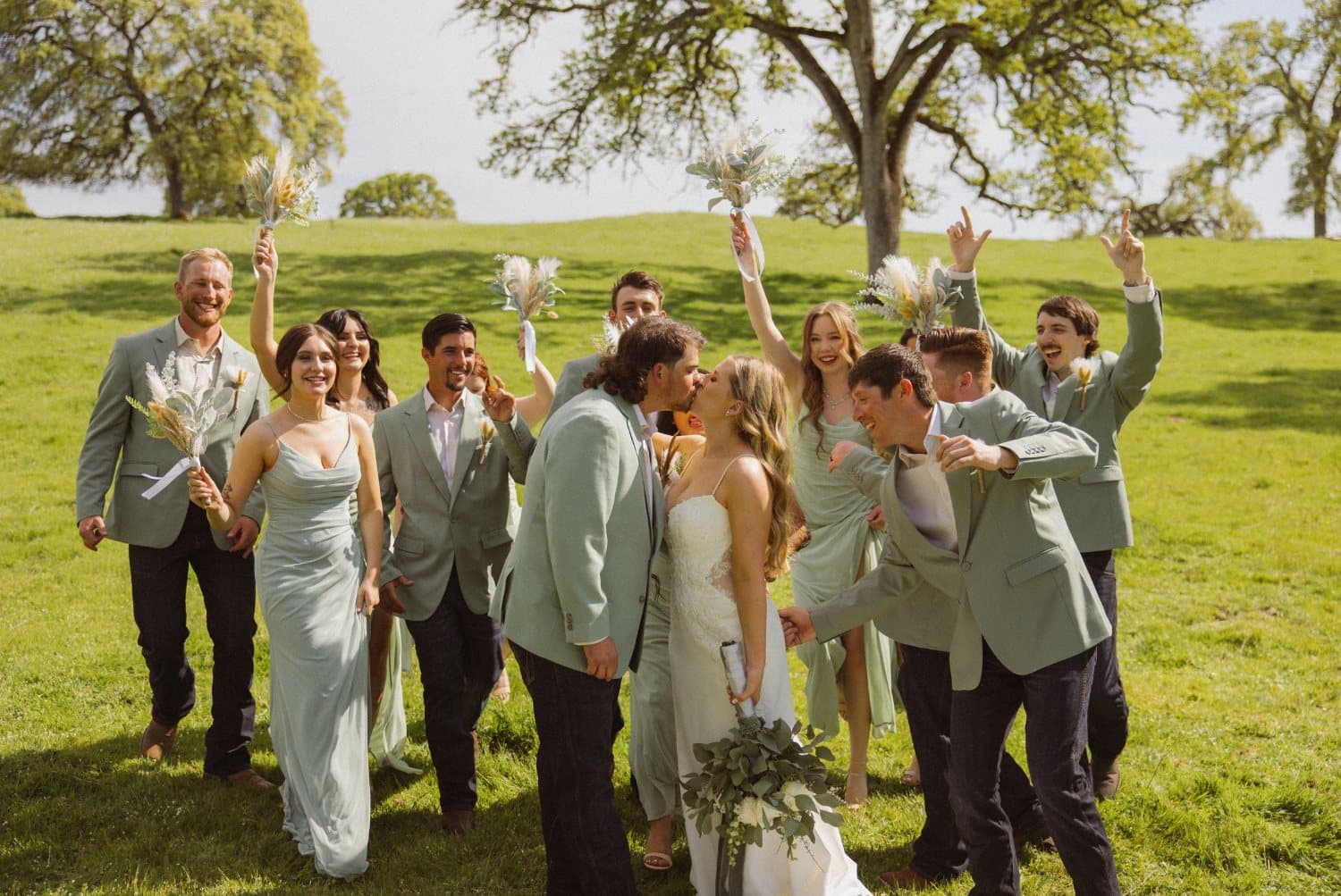 Bridesmaids in sage gowns walk arm in arm with the bride through the green oak meadow
