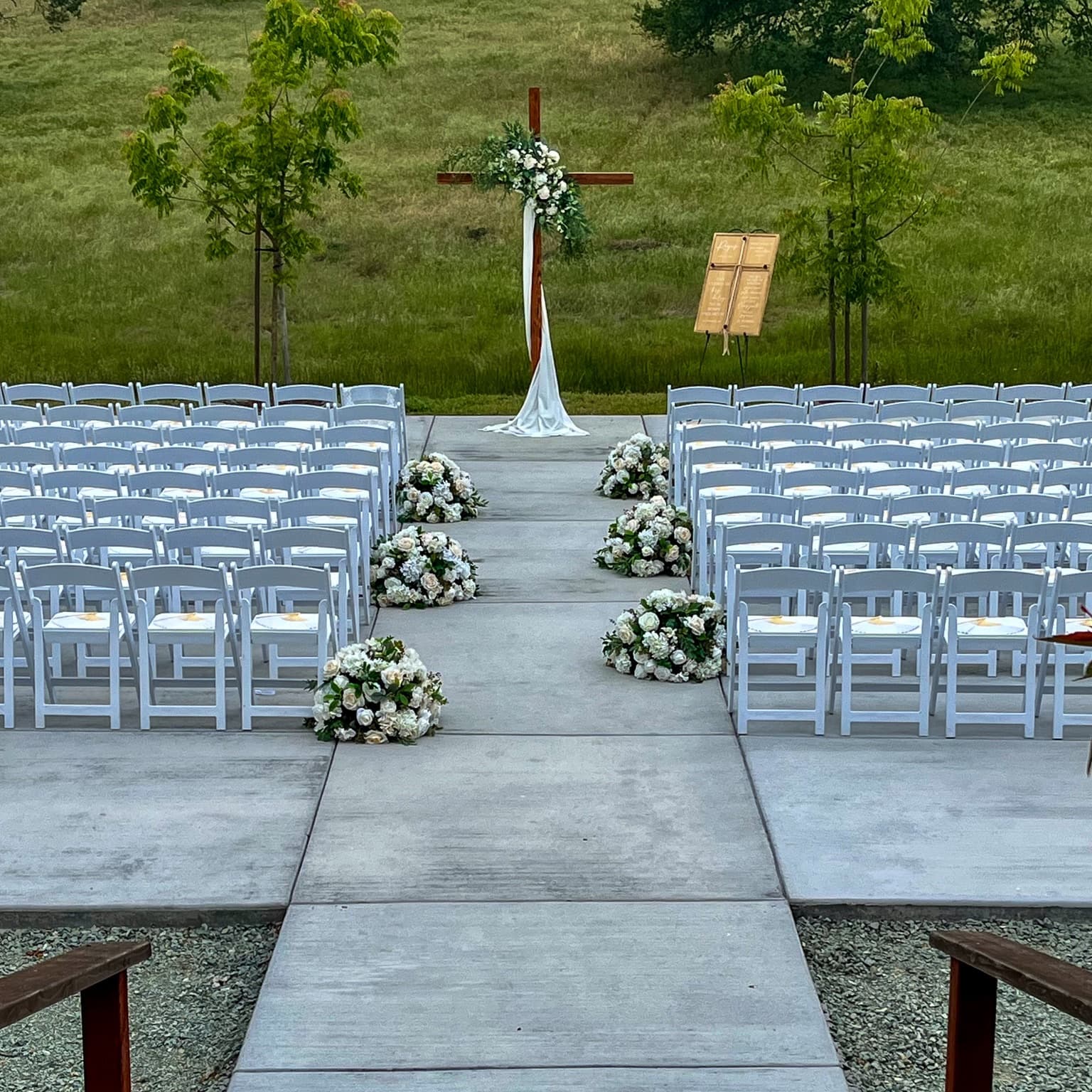 Ceremony aisle with white chairs and rose arrangements leading to a draped wooden cross against the green foothills near Plymouth, CA