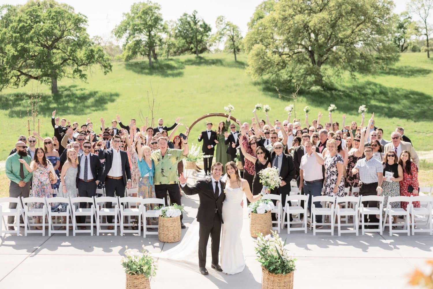 Newly married couple raise their arms as more than a hundred guests cheer from white chairs at the ranch ceremony site