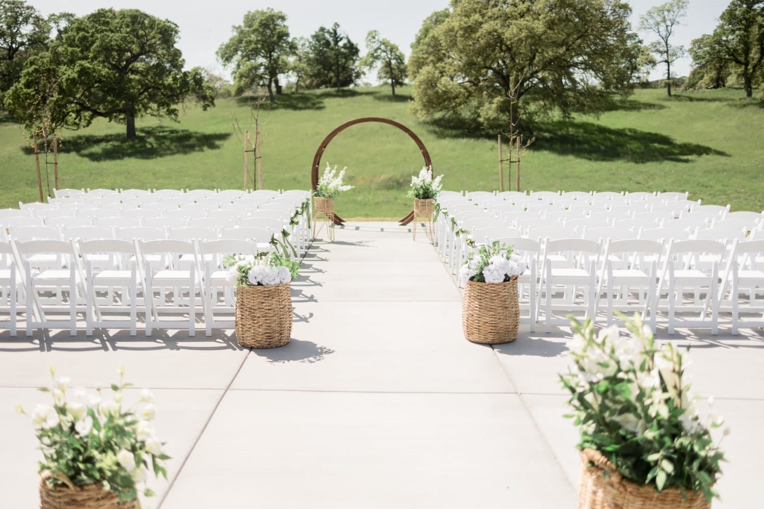 Ceremony aisle set with white folding chairs, woven baskets of white florals and a wooden circle arch on the oak-shaded ranch ceremony site