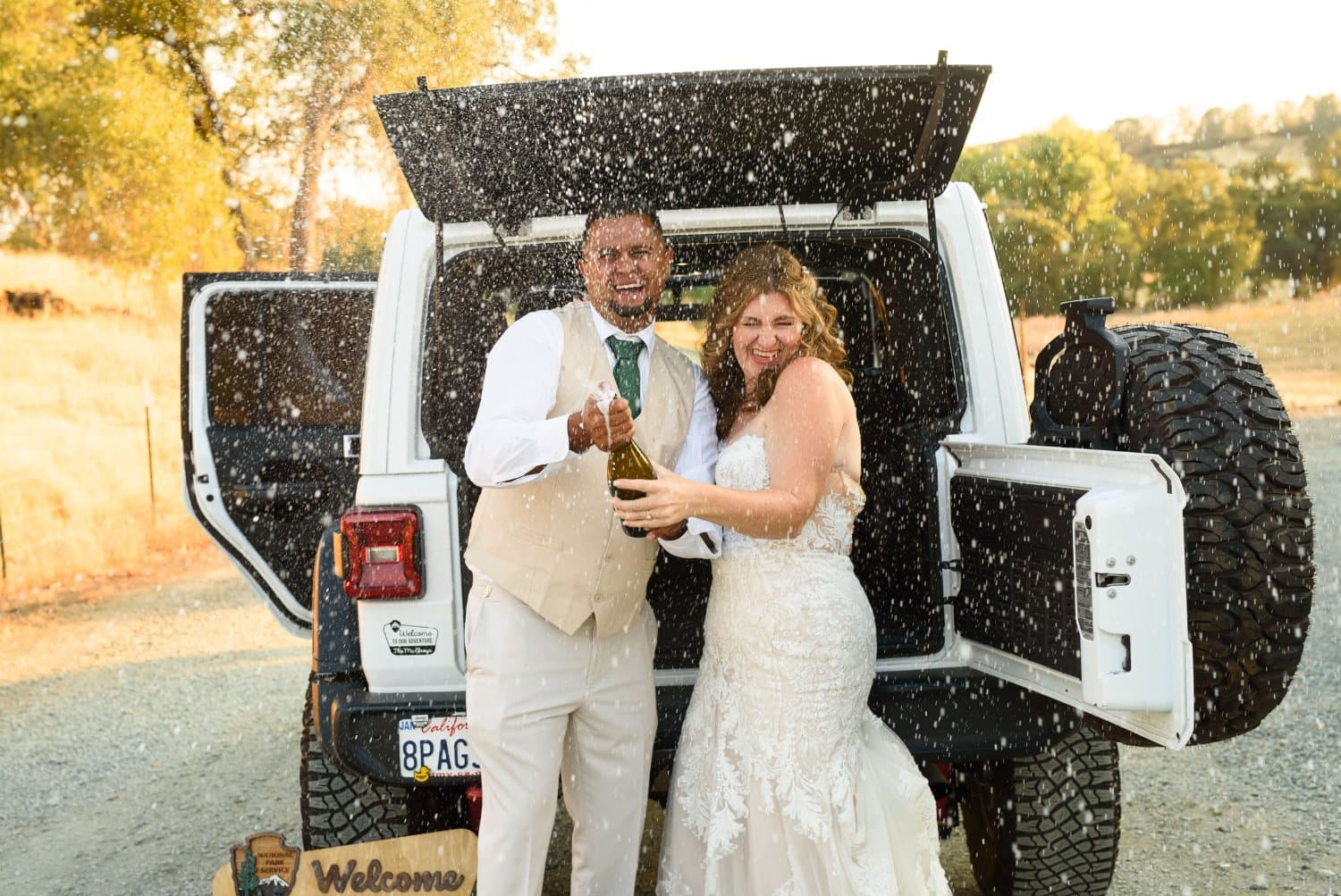 Couple laugh through a spray of champagne at the open tailgate of a white Jeep as the sun drops behind the hills