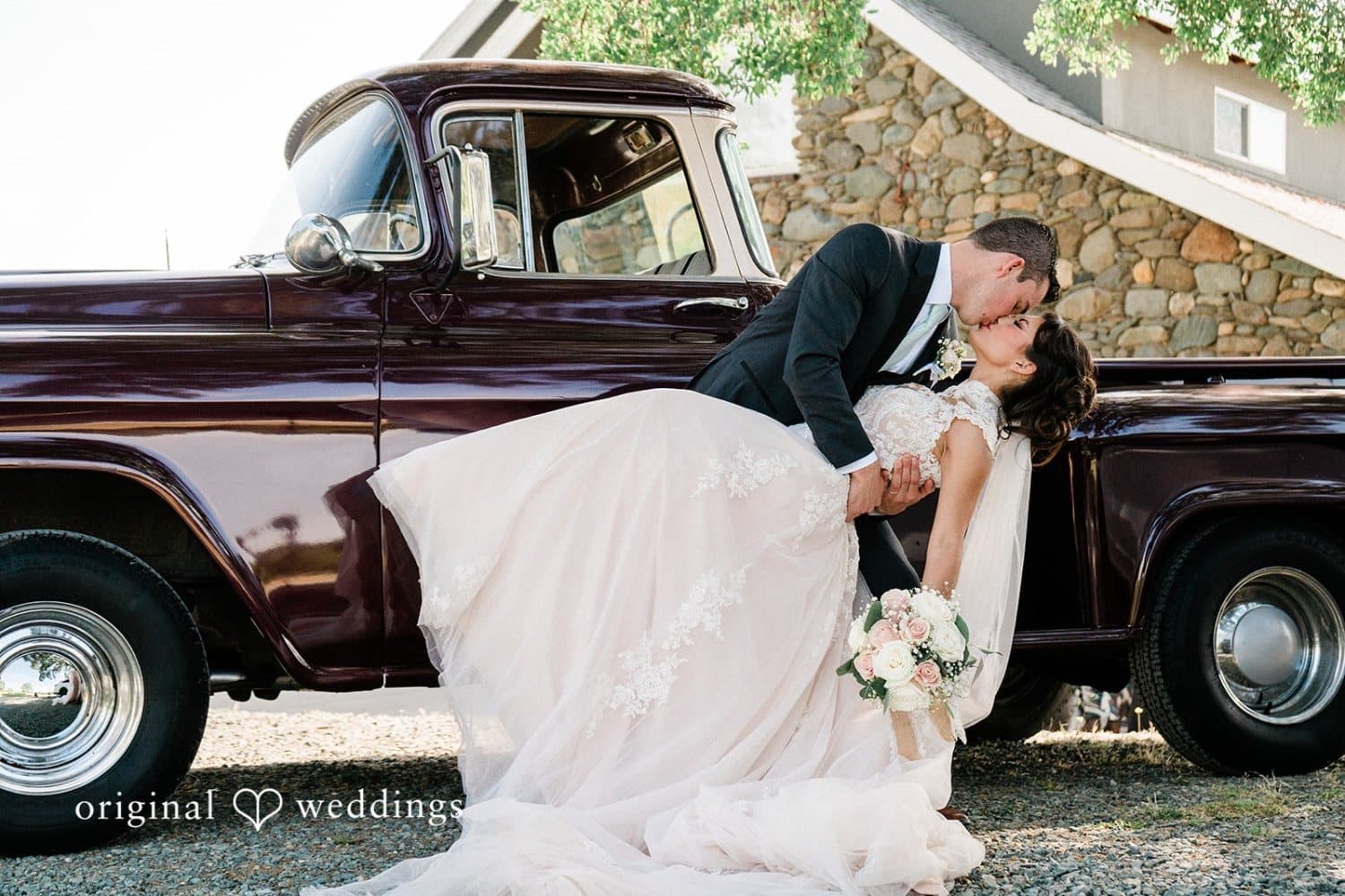 Groom dips the bride for a kiss against a maroon vintage pickup parked beside the river-rock barn