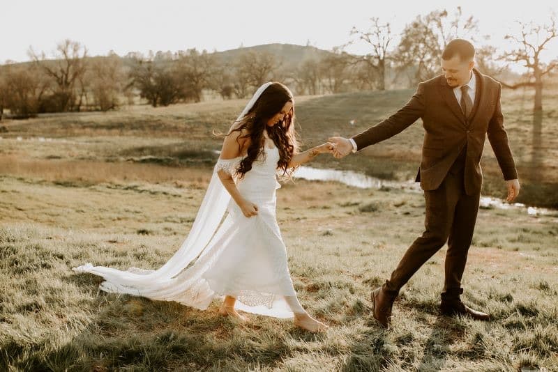 Groom leads the bride across the meadow at golden hour, the stock pond and valley oaks behind them