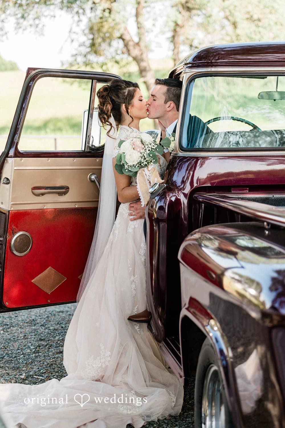 Bride and groom kiss in the open door of a maroon vintage truck with her bouquet of blush roses