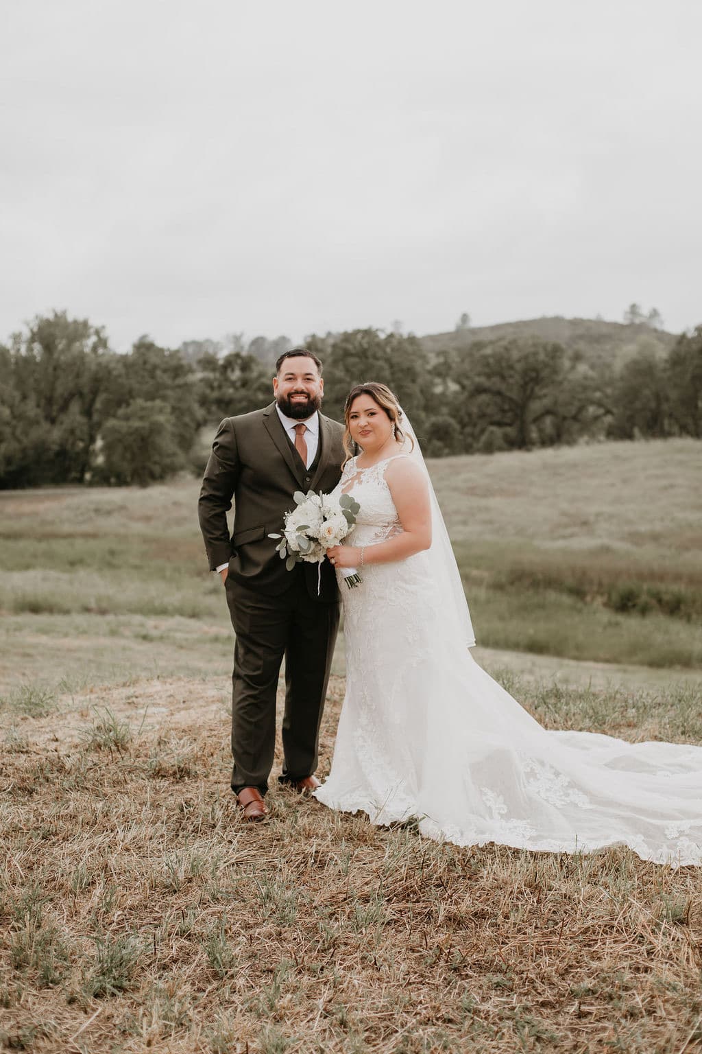 Bride and groom in an olive suit stand together in the oak meadow, her lace train sweeping the grass