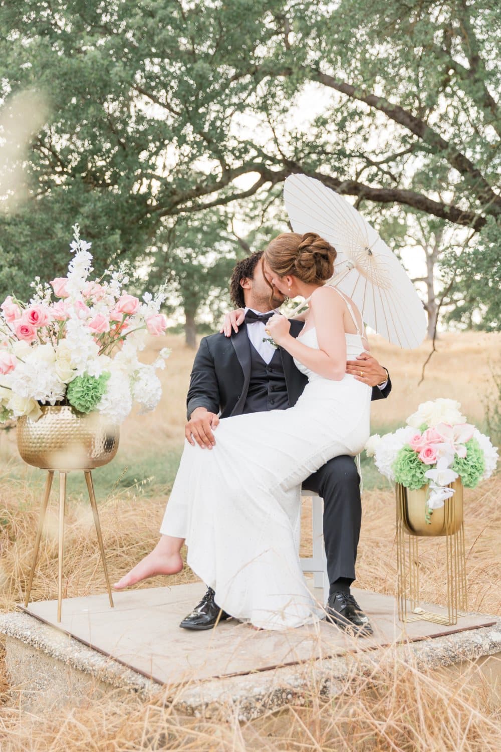 Couple kiss behind a paper parasol on a stone pad in the dry grass, flanked by brass stands of pink and white florals