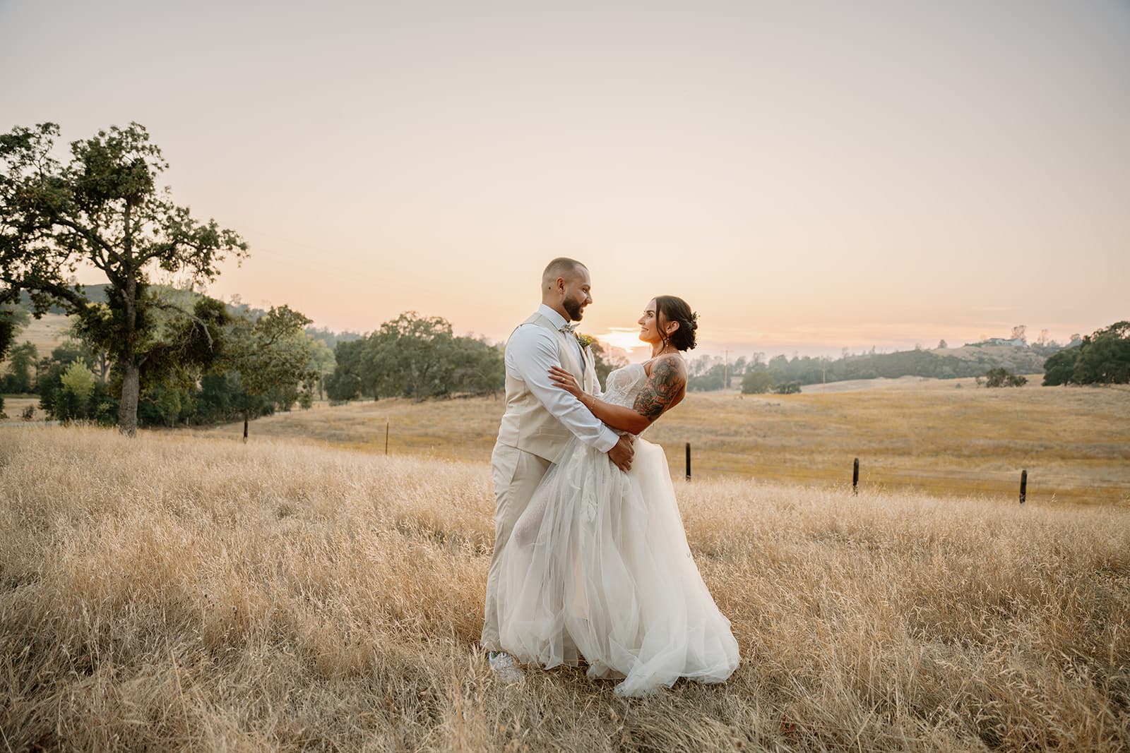 Couple embrace in the dry September grass at sunset along the ranch fence line, country wedding in Amador County