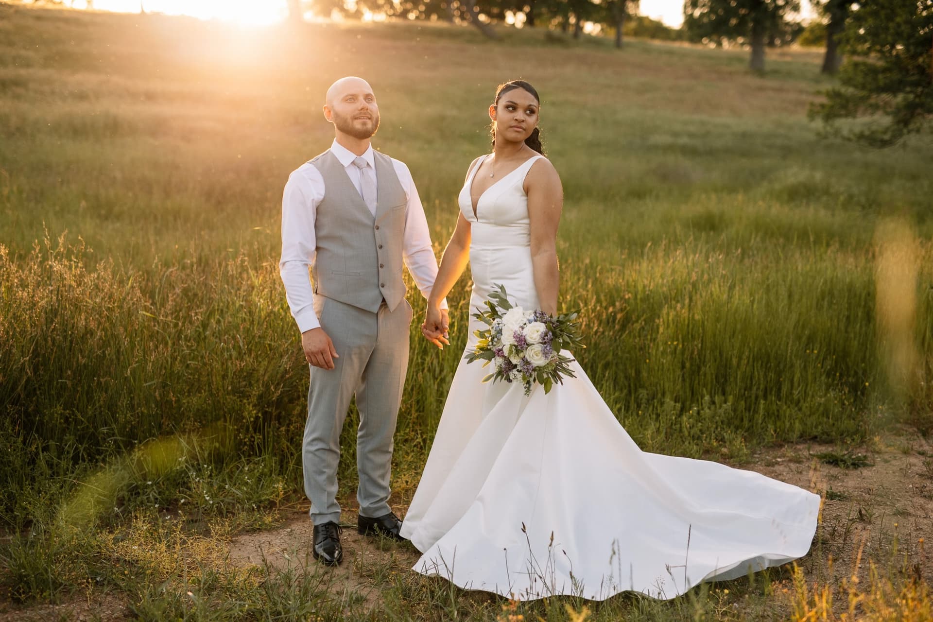 Bride and groom hold hands in waist-high green grass as the setting sun flares over the hill, Sierra foothills wedding