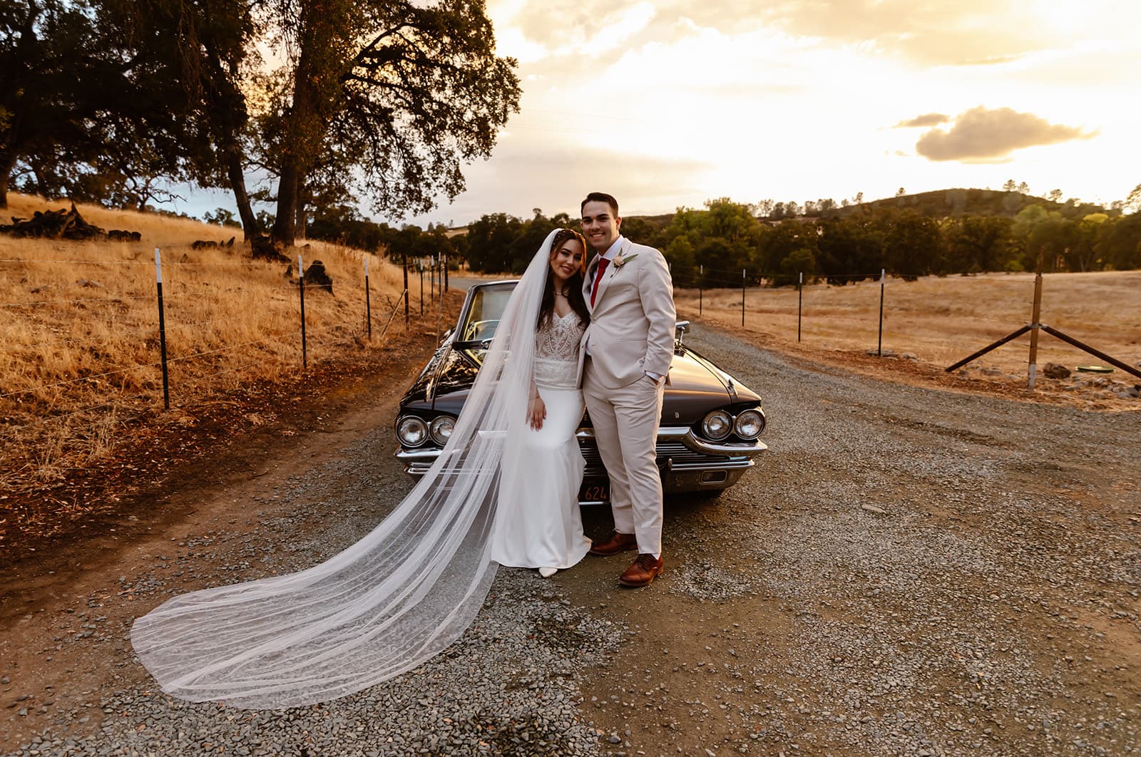 Newlyweds lean on a vintage black coupe on the gravel ranch road, her cathedral veil trailing to the ground