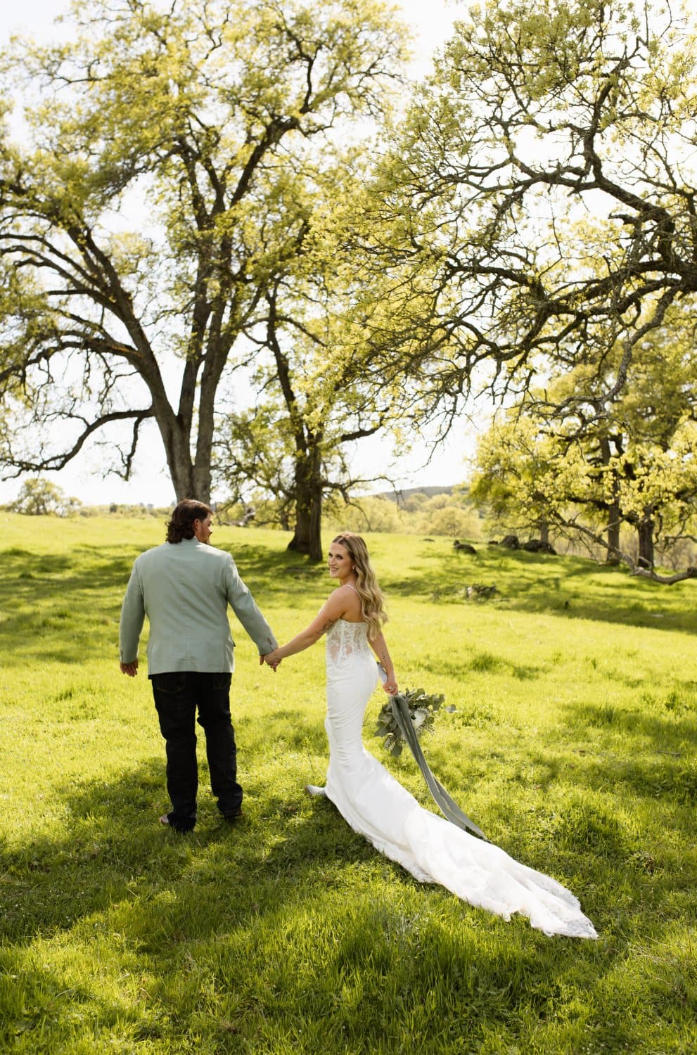 Bride looks back over her shoulder while walking hand in hand with the groom through the green oak meadow