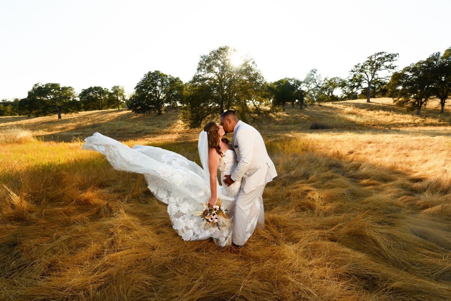 Couple kiss in waist-high golden grass as the bride's lace train sweeps behind her in the low evening sun