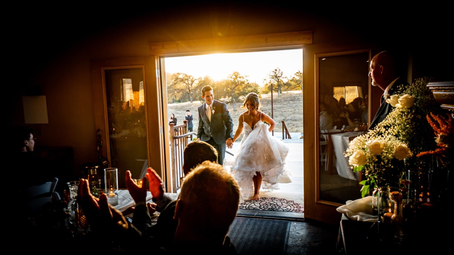 Newlyweds enter the reception through the open barn doors at sunset as seated guests applaud