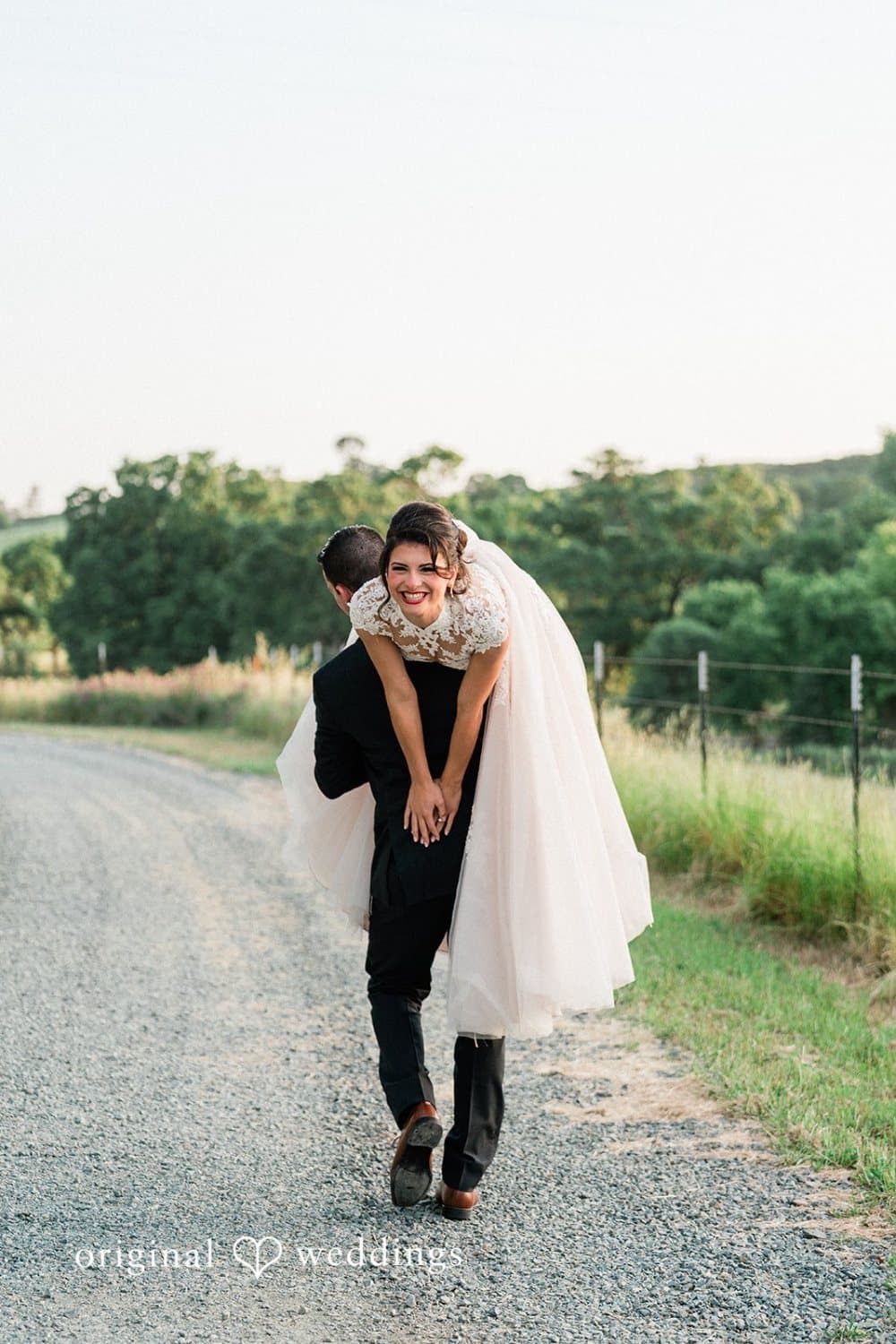 Groom carries the laughing bride piggyback down the gravel ranch road at dusk