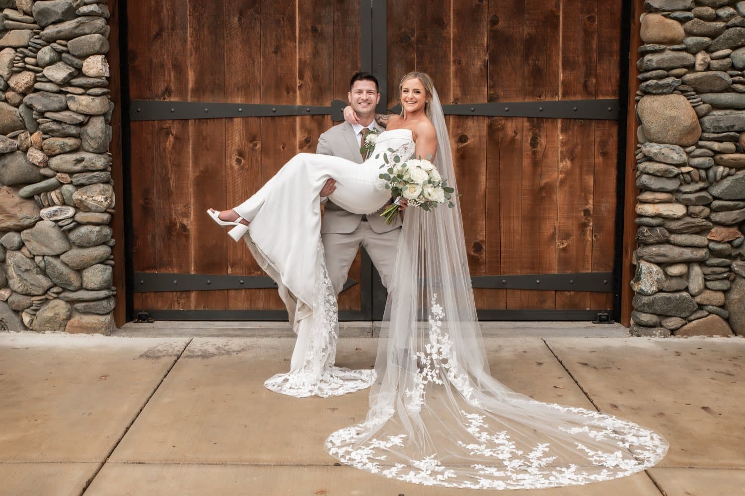 Groom lifts the bride off her feet in front of the stone barn doors, her lace veil pooled on the concrete