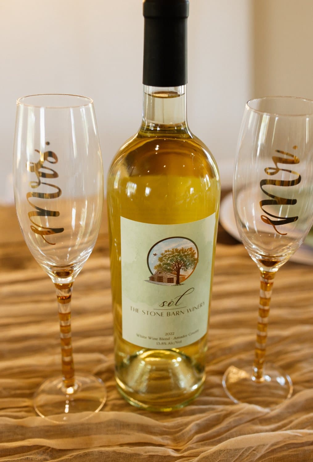 Bottle of sēl white wine from The Stone Barn Winery between gold Mr and Mrs champagne flutes on a cheesecloth runner