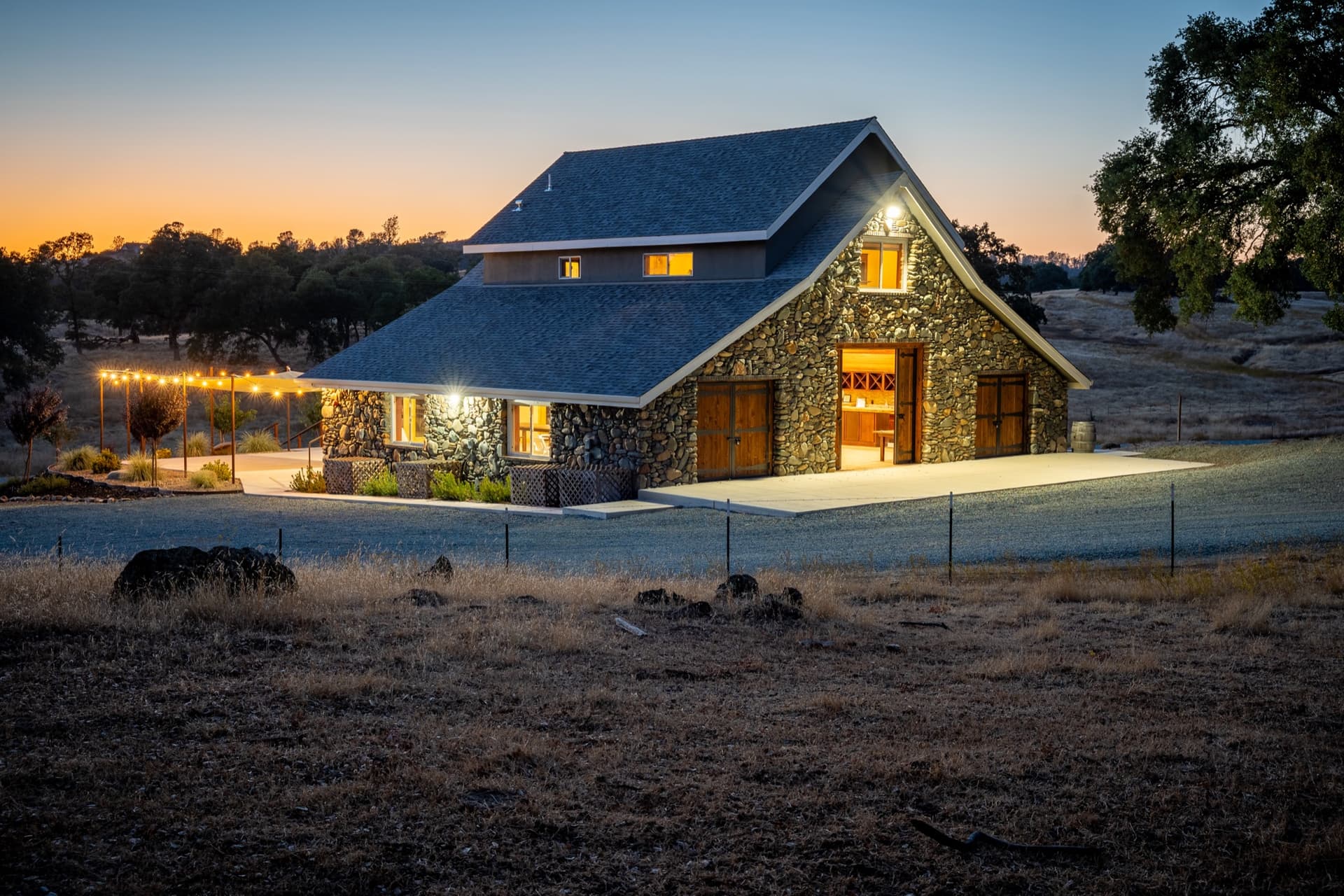 Barn wedding venue in the Sierra foothills at sunset: the stone barn across the dry field, string lights and glowing doors