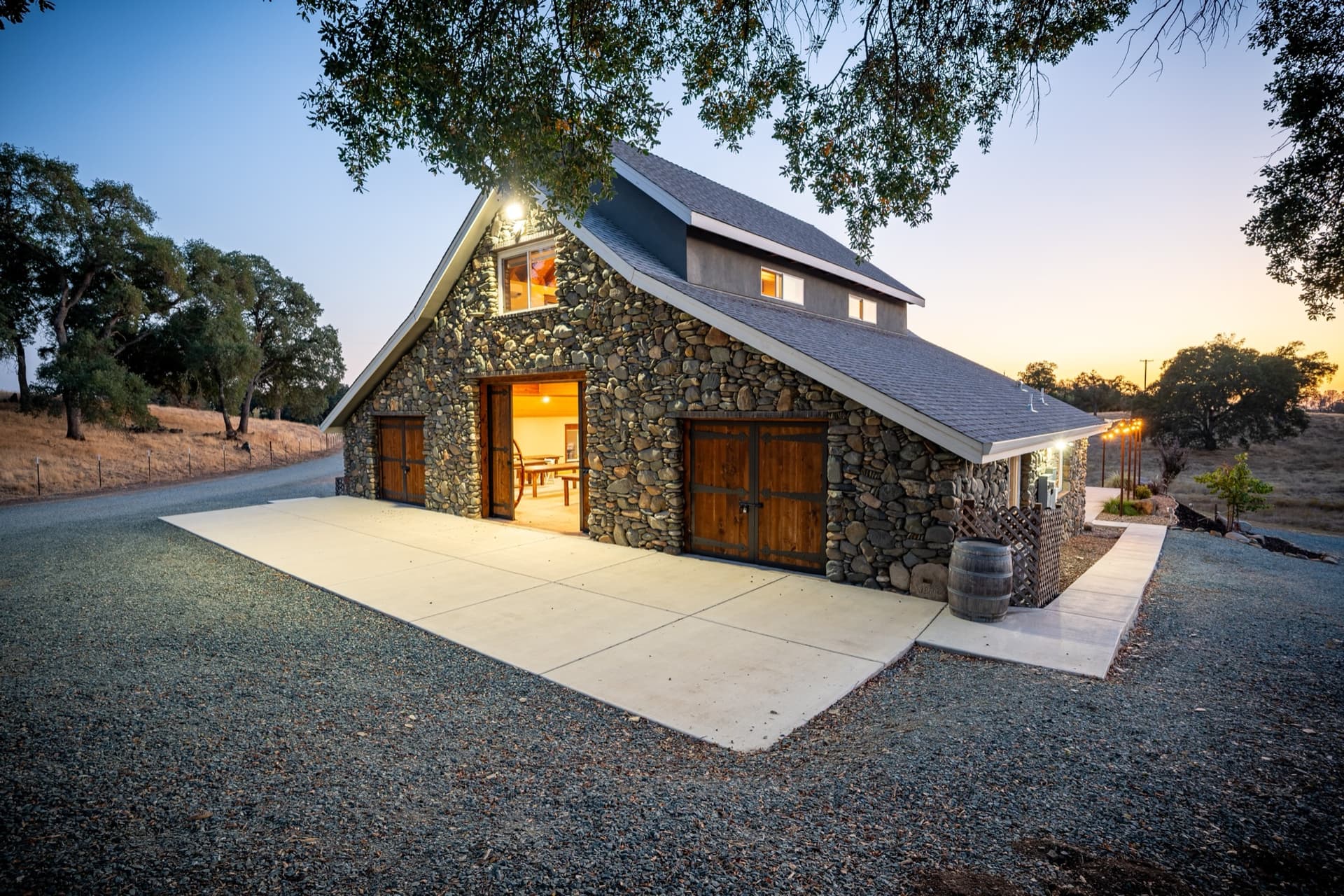 Rustic barn wedding reception at dusk: timber doors open onto the lit patio, a wine barrel at the corner, oaks overhead