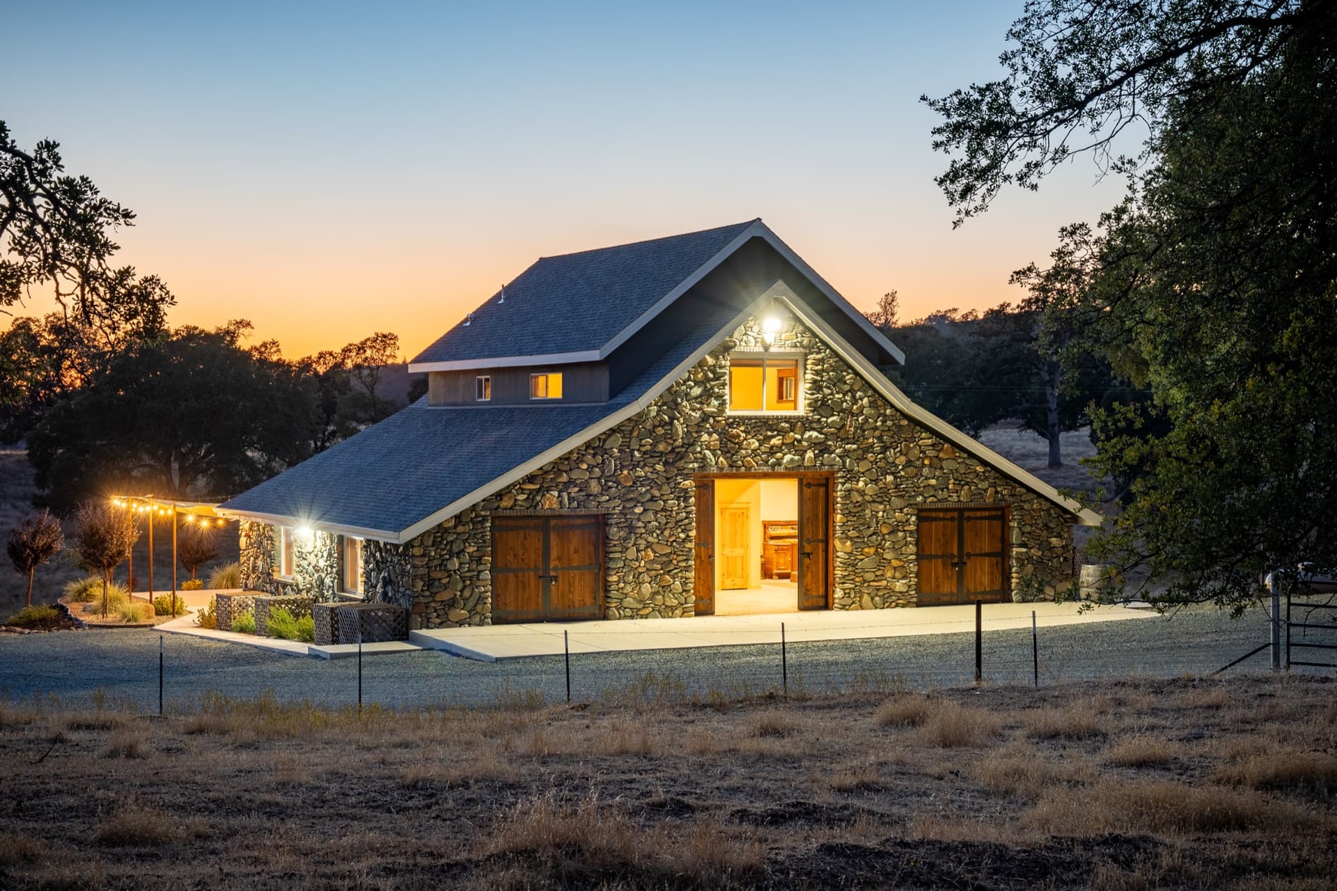 The stone barn head-on after sunset with the centre doors open on a candlelit reception, orange sky behind the oaks