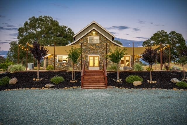 Front of the stone barn at blue hour with shade sails, redwood steps and planted beds along the gravel drive