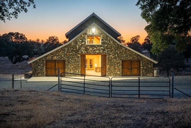 The barn seen across the ranch gate after sunset, windows glowing under a peach sky