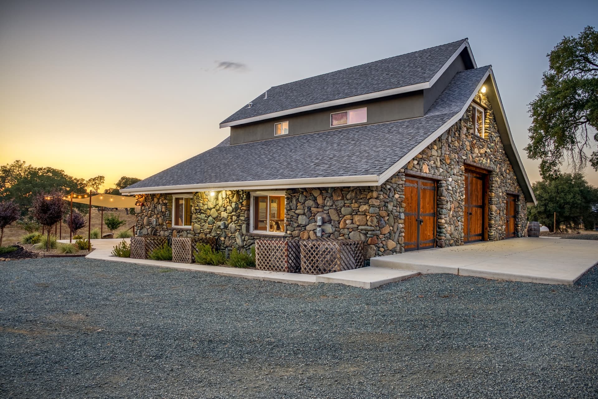 Country wedding venue at sunset: string lights and a shade sail over the stone barn's patio, lattice planters below the windows