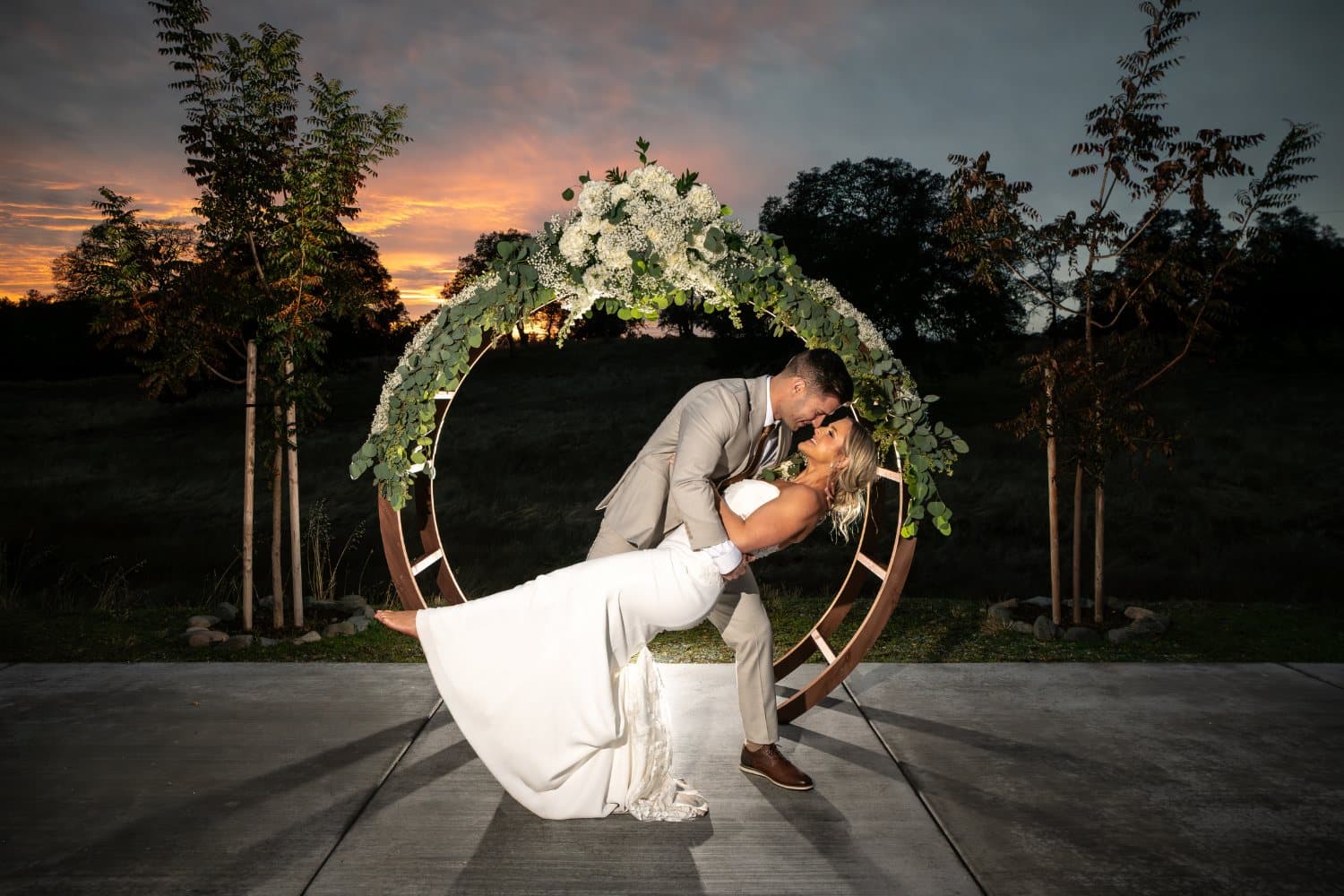Bride and groom laugh under the eucalyptus-and-rose circle arch as the sky turns orange behind the ranch oaks