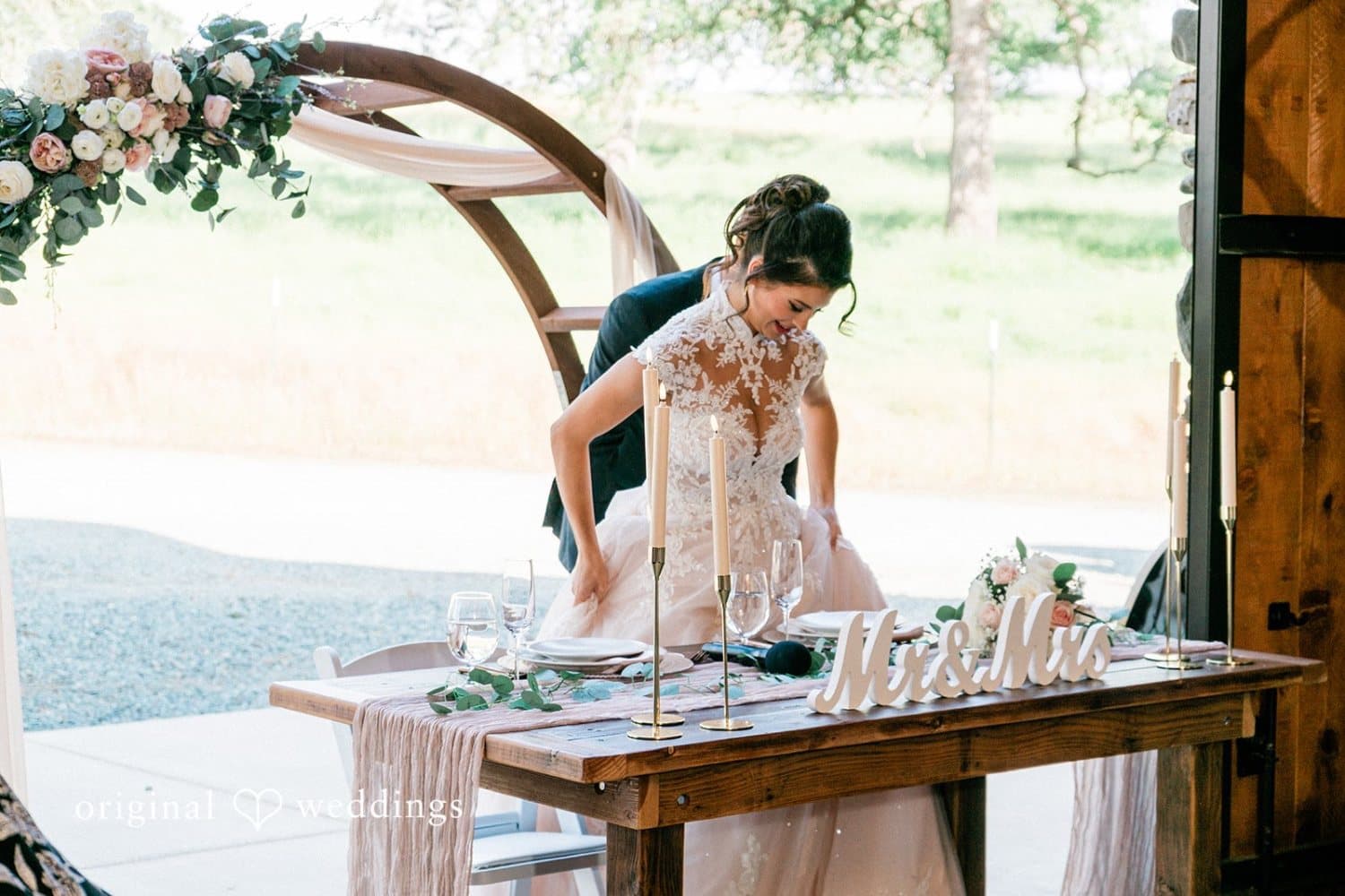 Sweetheart table with taper candles and a Mr and Mrs sign in the stone barn doorway, framed by a floral arch