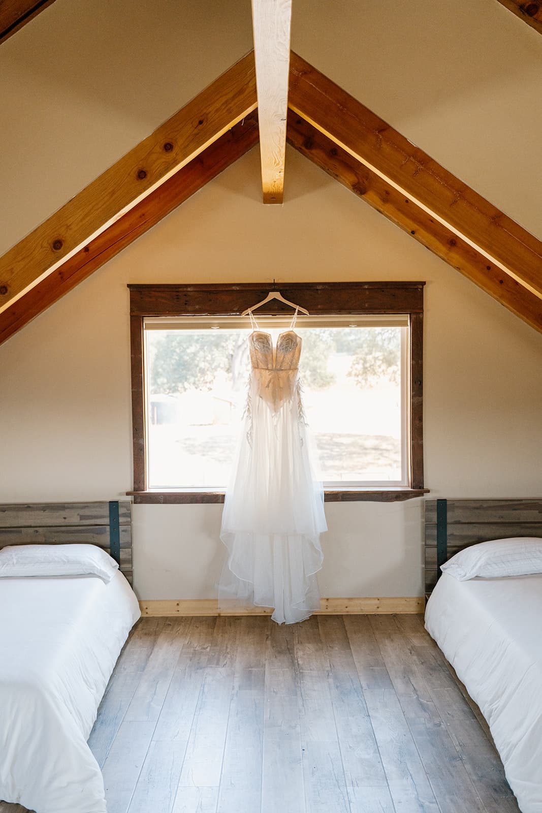 Wedding gown hanging in the barn loft window between exposed timber beams, backlit by morning light