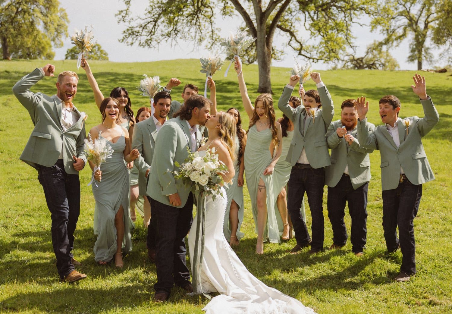 Wedding party in sage jackets cheer around the couple as they kiss in the spring meadow