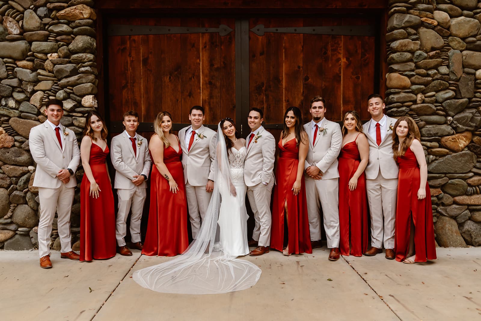 Wedding party in red satin gowns and tan suits at the stone barn's timber doors, the bride's veil pooled on the concrete