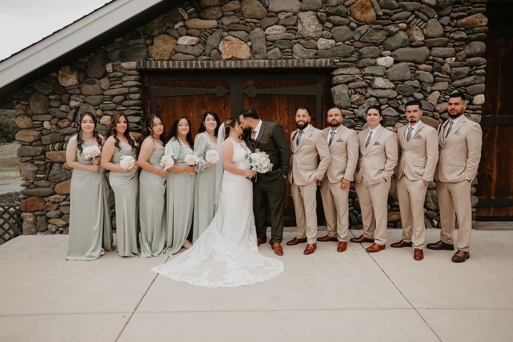 Couple kiss between sage-dressed bridesmaids and tan-suited groomsmen at the river-rock barn doors, rustic barn wedding