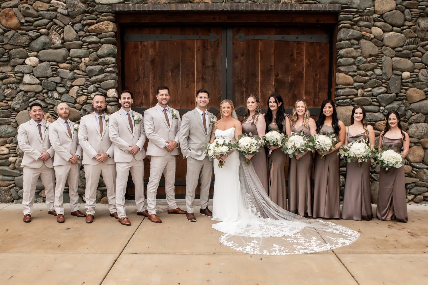 Full wedding party in tan suits and mocha satin gowns lined up in front of the stone barn's wooden doors