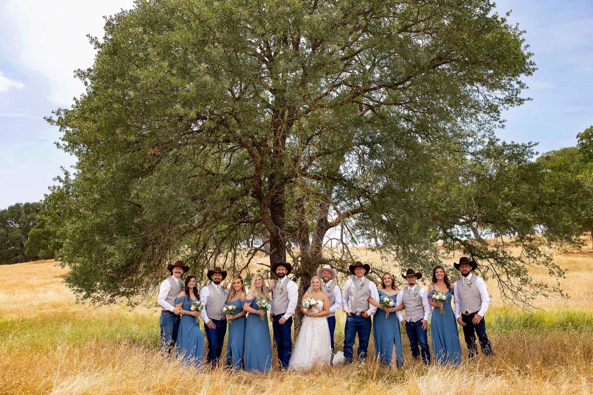 Western wedding party in cowboy hats, dusty-blue gowns and denim beneath an oak in the golden grass, country wedding in California